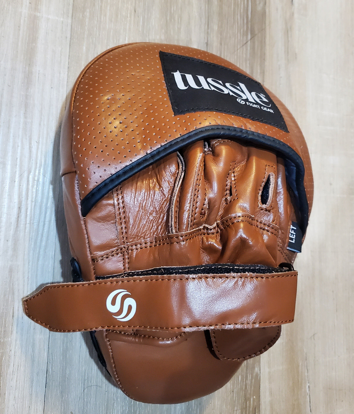 A brown leather martial arts focus mitt with a black and white 'Lussie' brand patch and a white circular logo on the strap, resting on a light wood floor.